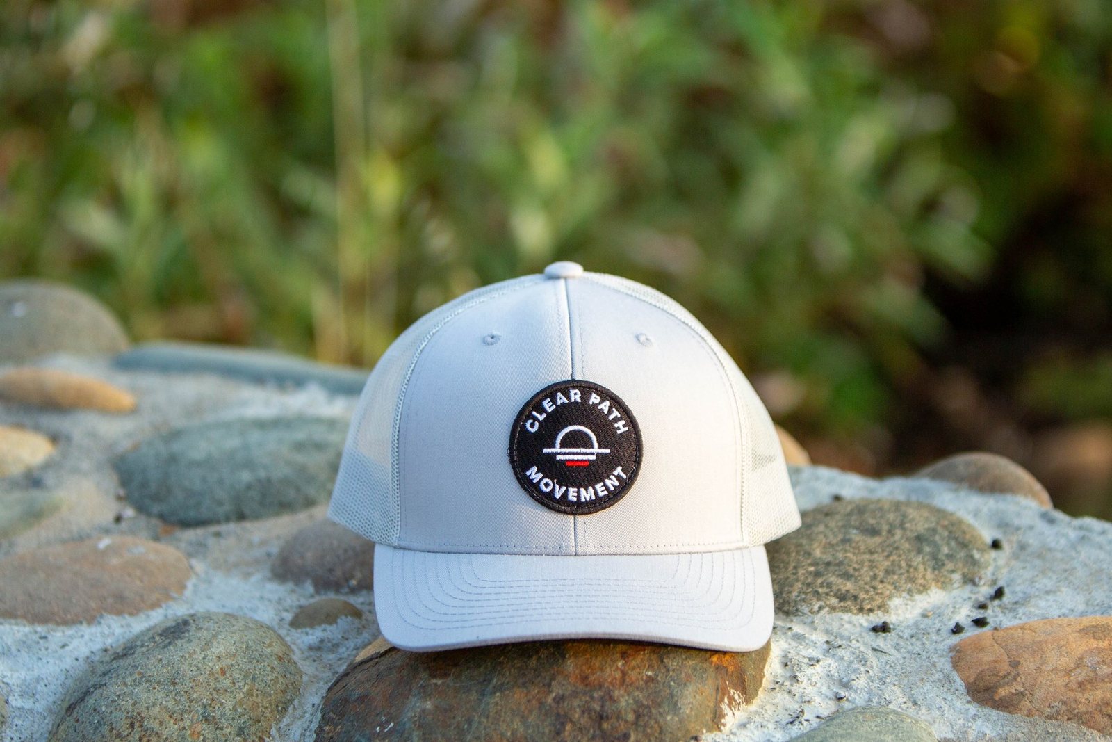 Grey Clear Path Movement trucker hat resting on river stones