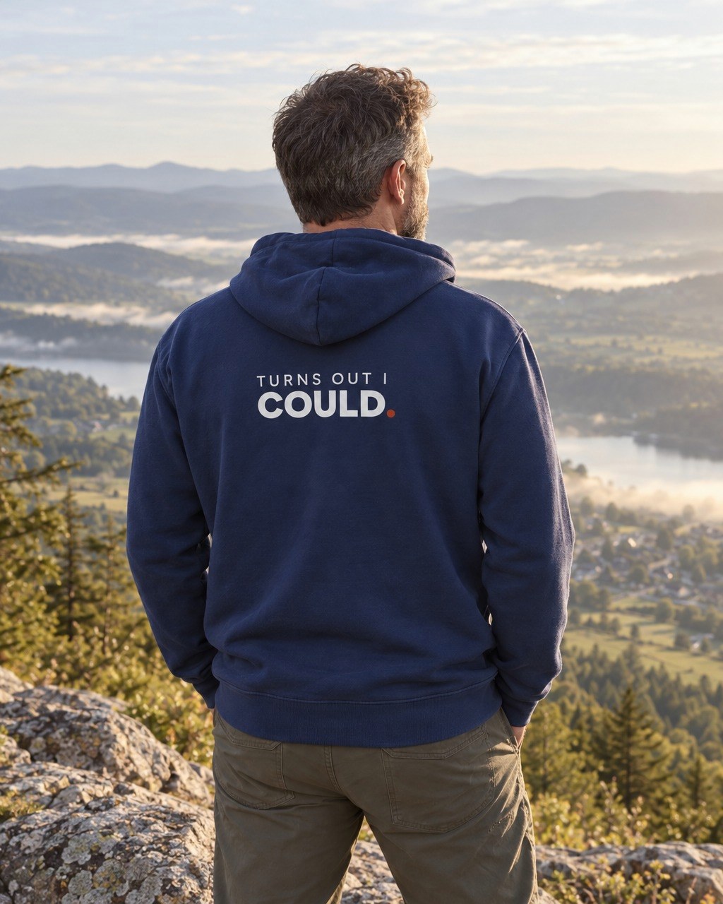 Man on a mountain overlook wearing the navy Turns Out I Could hoodie, back print showing